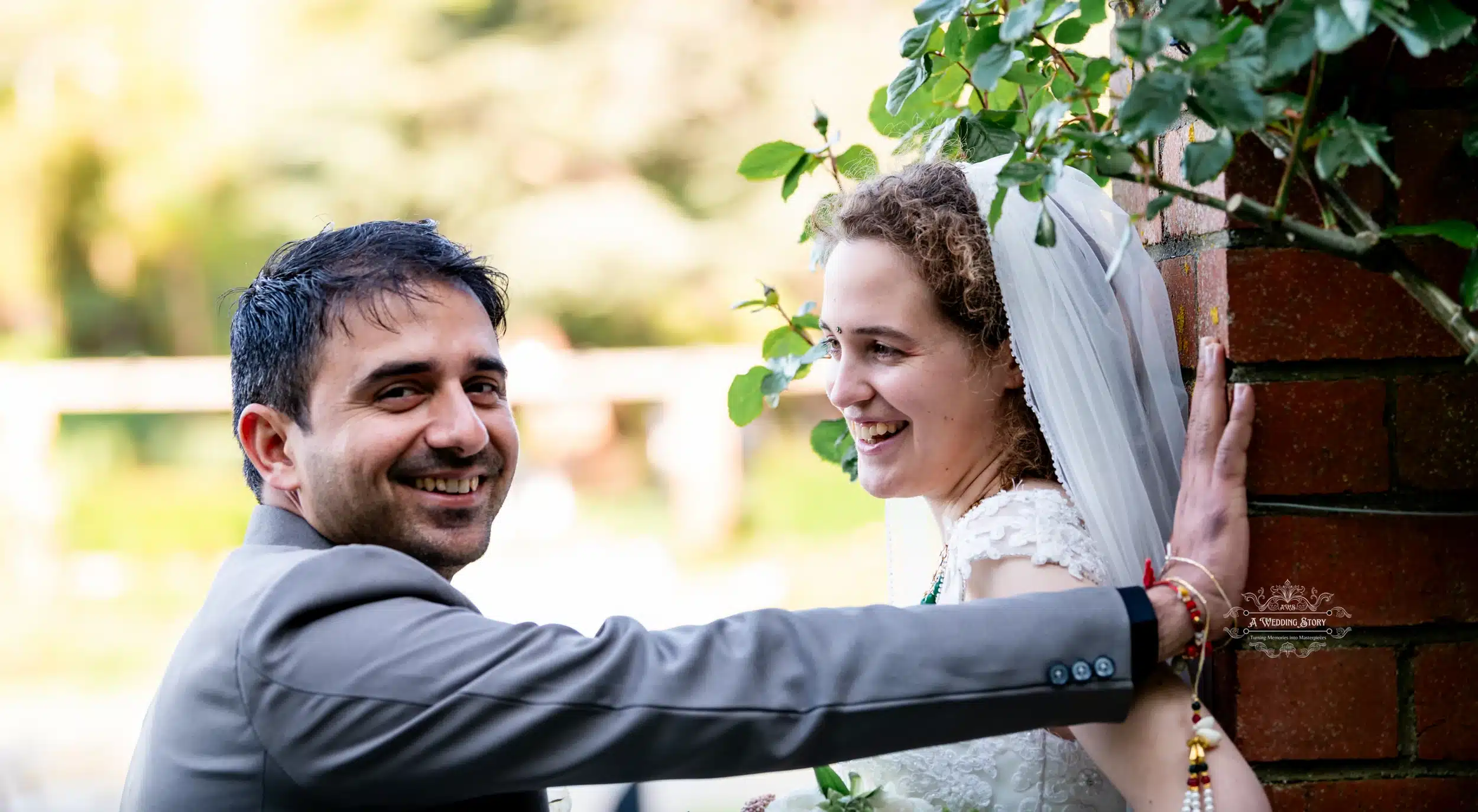 Groom smiling at the camera while leaning on a brick wall, with the bride in a veil looking at him, both sharing a joyful moment