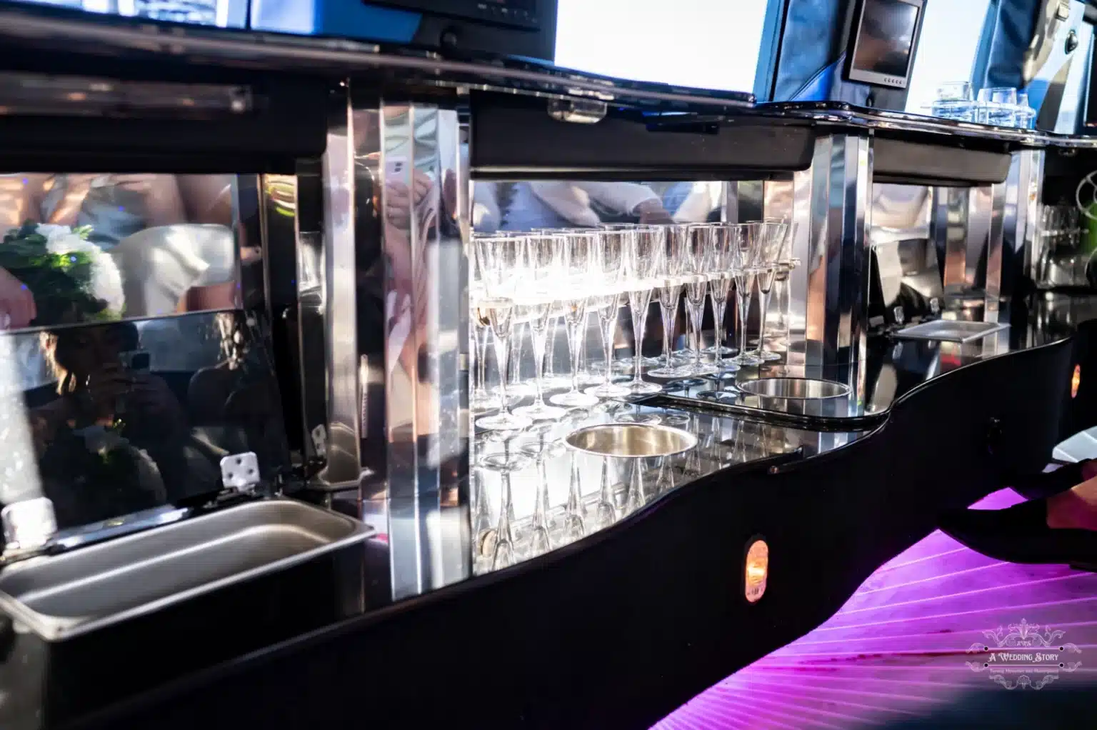 A sophisticated bar setup inside a luxury limousine featuring champagne flutes neatly arranged with a reflective design, illuminated with ambient purple lighting for an elegant experience.