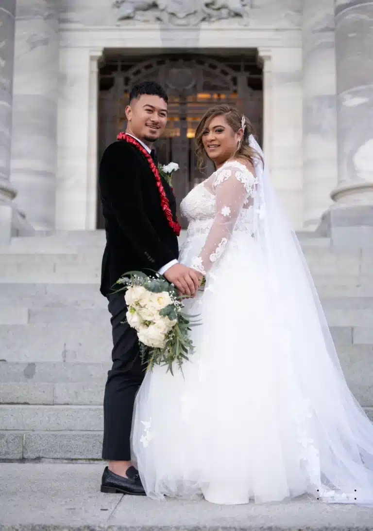 Bride and groom smiling during a wedding photoshoot at a historic location in Wellington, New Zealand, holding a bouquet of white flowers.