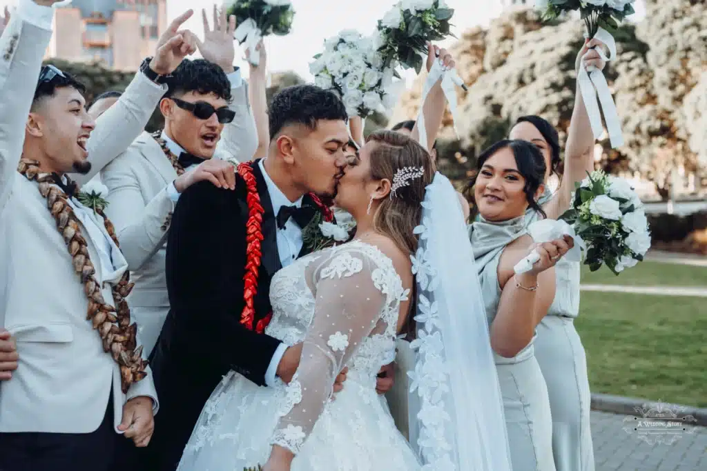 Bride and groom share a romantic kiss surrounded by their bridal party during a vibrant wedding celebration in Wellington, New Zealand.