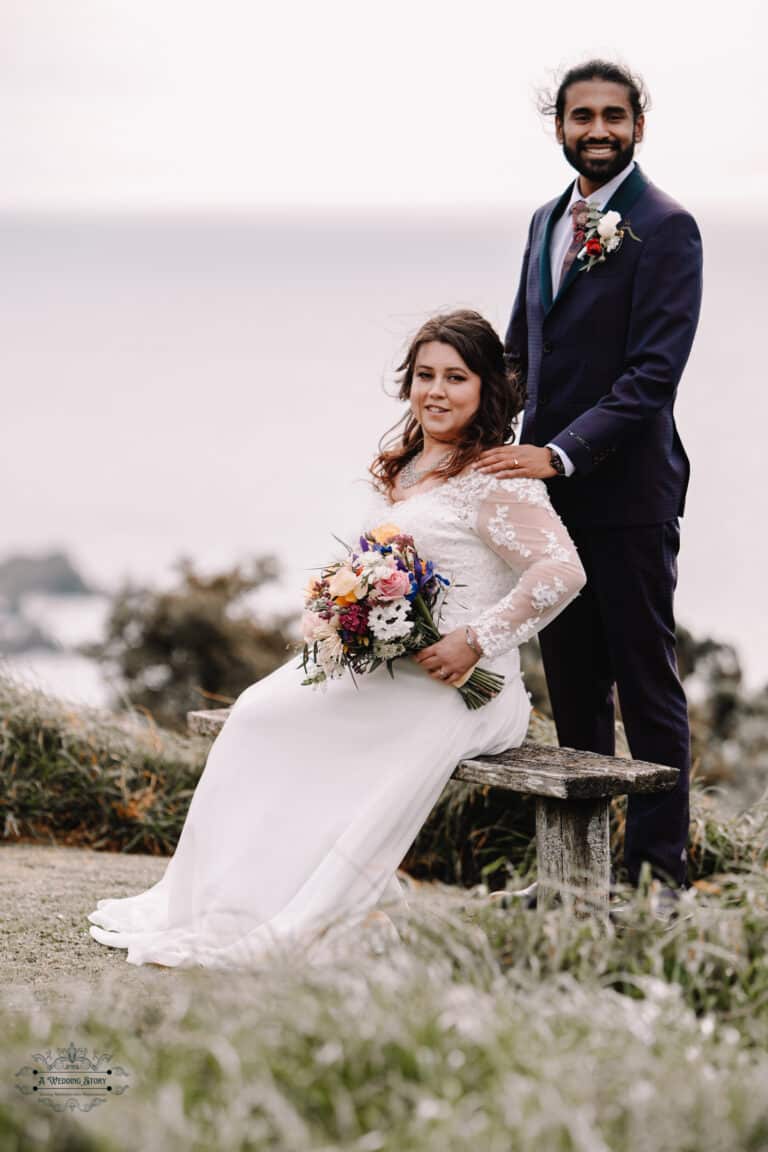 A bride holding a colorful bouquet seated on a bench while the groom stands behind her during their Boomrock wedding in Wellington.