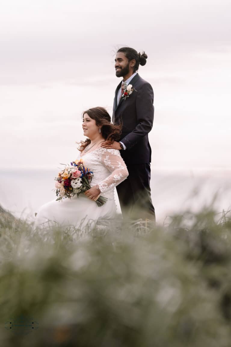 A bride holding a vibrant bouquet while the groom stands behind her on a scenic hillside at Boomrock, Wellington.