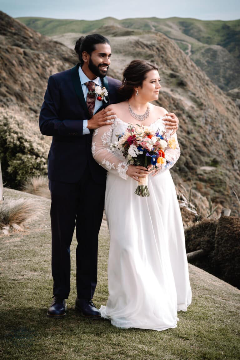 A couple standing together on a scenic hill at Boomrock, Wellington, holding a vibrant bridal bouquet.