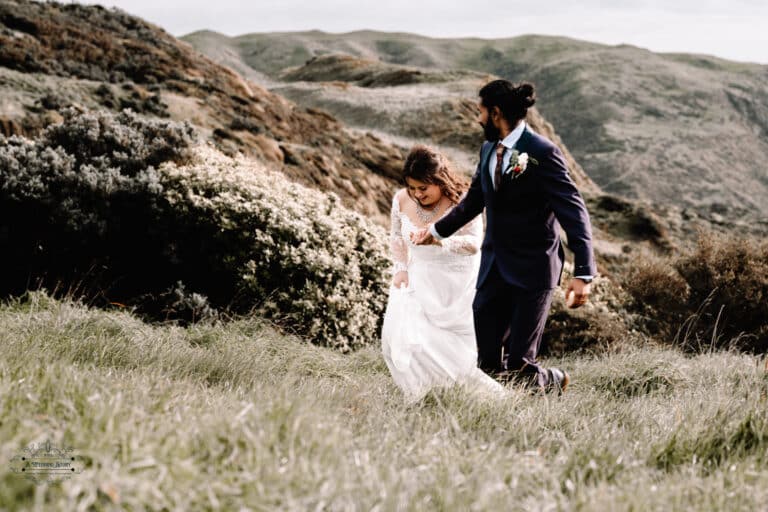 A couple walking hand-in-hand through lush grass on a scenic hillside at Boomrock, Wellington, during their wedding day.