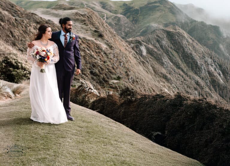 A newlywed couple walking along the dramatic cliffs of Boomrock, Wellington, with breathtaking landscapes in the background.