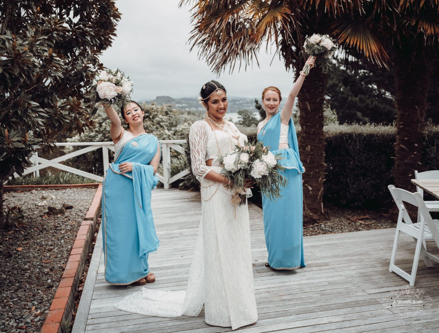 Bride in an elegant white gown holding a bouquet, joined by two bridesmaids in blue sarees, joyfully celebrating outdoors at The Lodge, Wellington