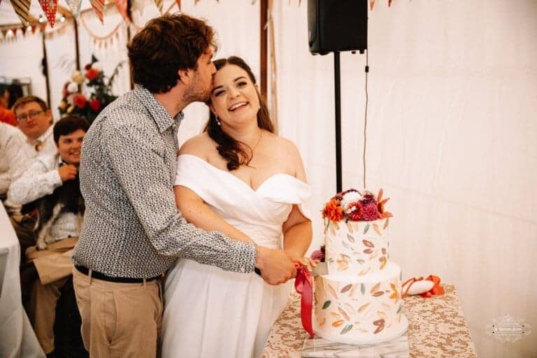 Groom kisses the bride’s forehead as they cut their wedding cake together in a festive reception setting