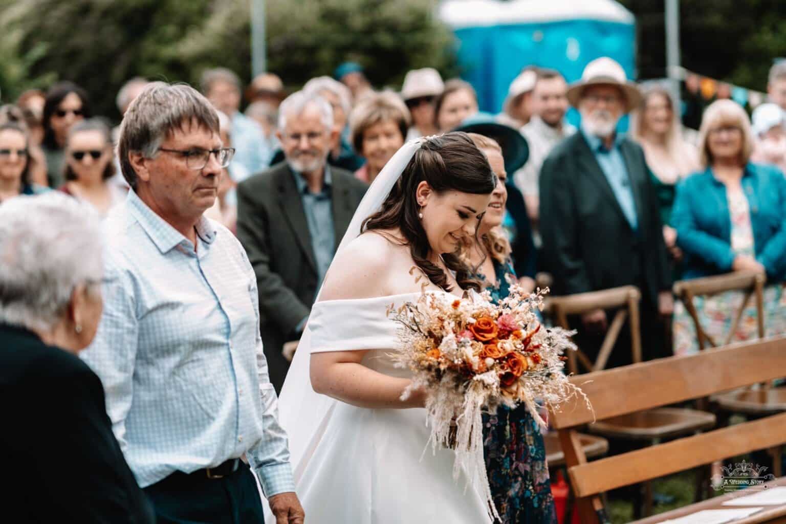 The bride walks down the aisle, holding a rustic bouquet, surrounded by family and friends