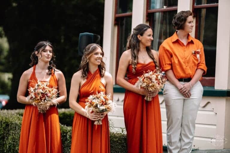 Bridesmaids in matching burnt-orange dresses holding floral bouquets, smiling during the wedding ceremony