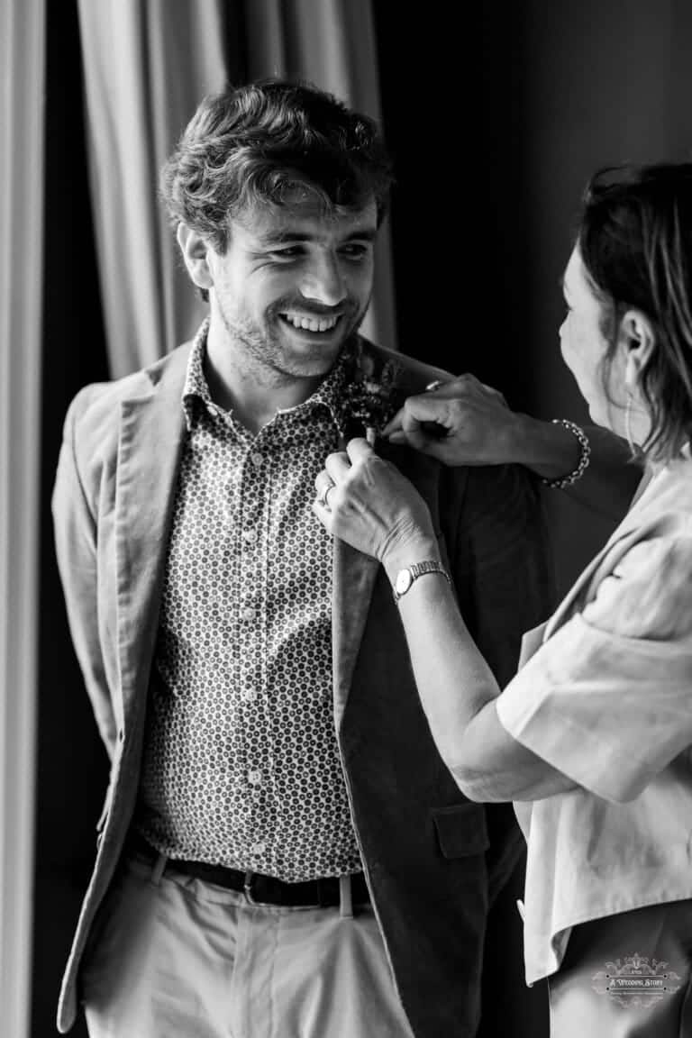 Black and white candid photo of the groom smiling while a loved one pins his boutonnière before the wedding in Carterton, New Zealand