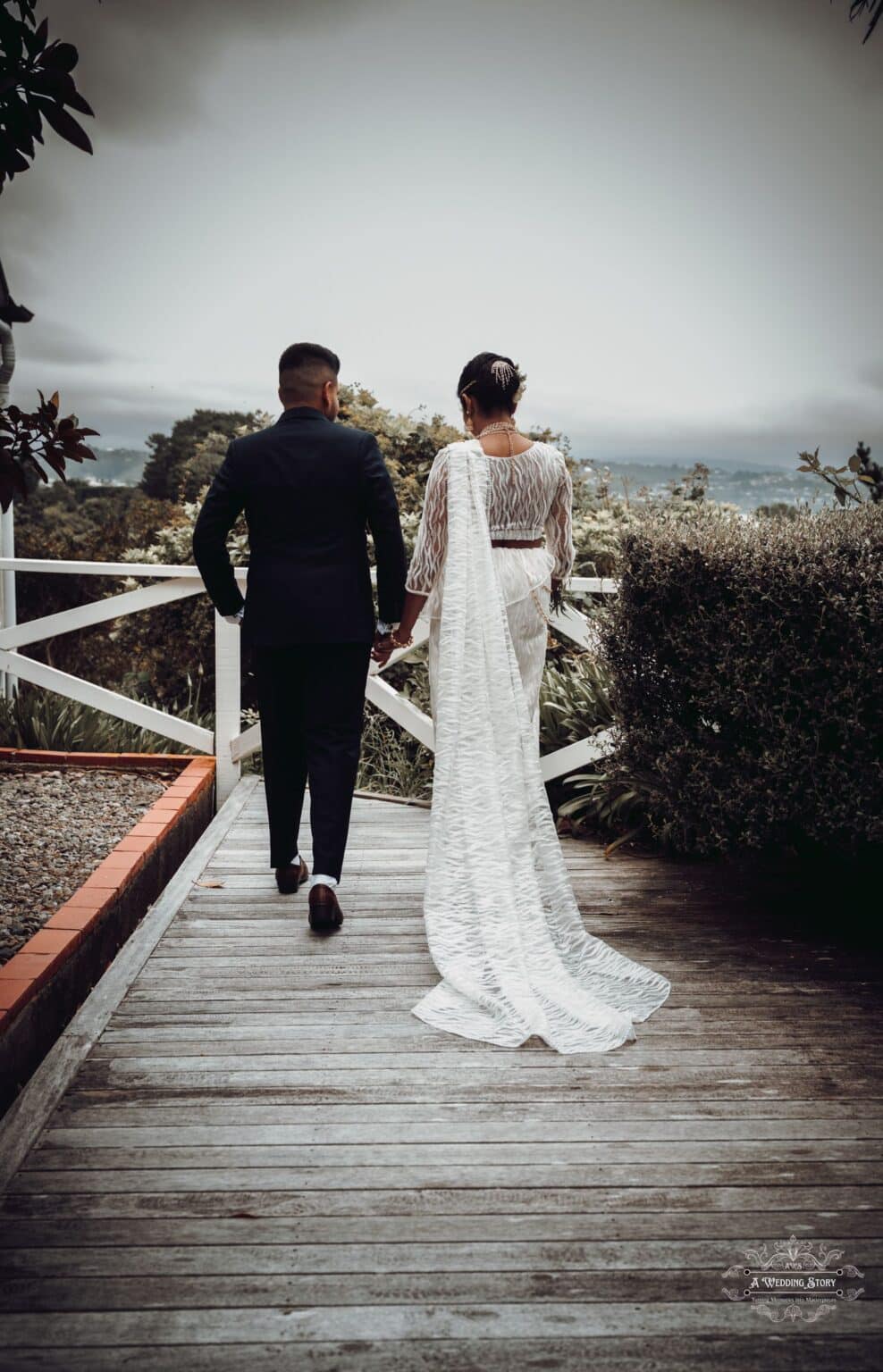 Kavi and Thisali walk hand in hand along a wooden pathway at The Lodge, Wellington, with stunning natural scenery in the background, symbolizing their journey together