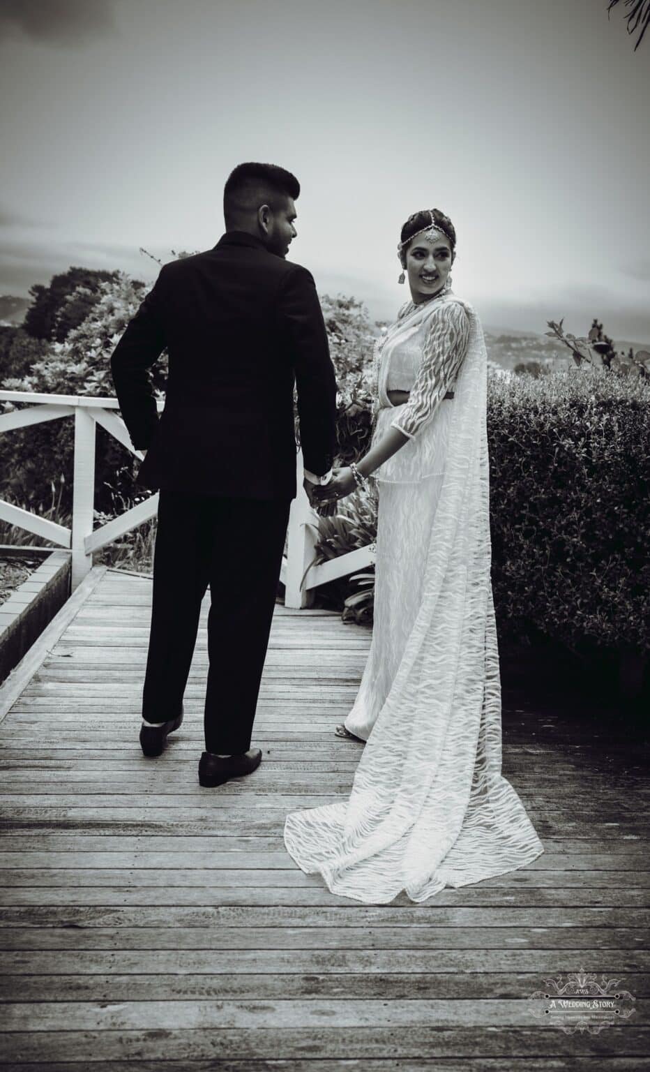 A newlywed couple walks hand in hand on a wooden pathway, with the bride in a traditional white saree looking back with a joyful expression, surrounded by lush greenery in Wellington