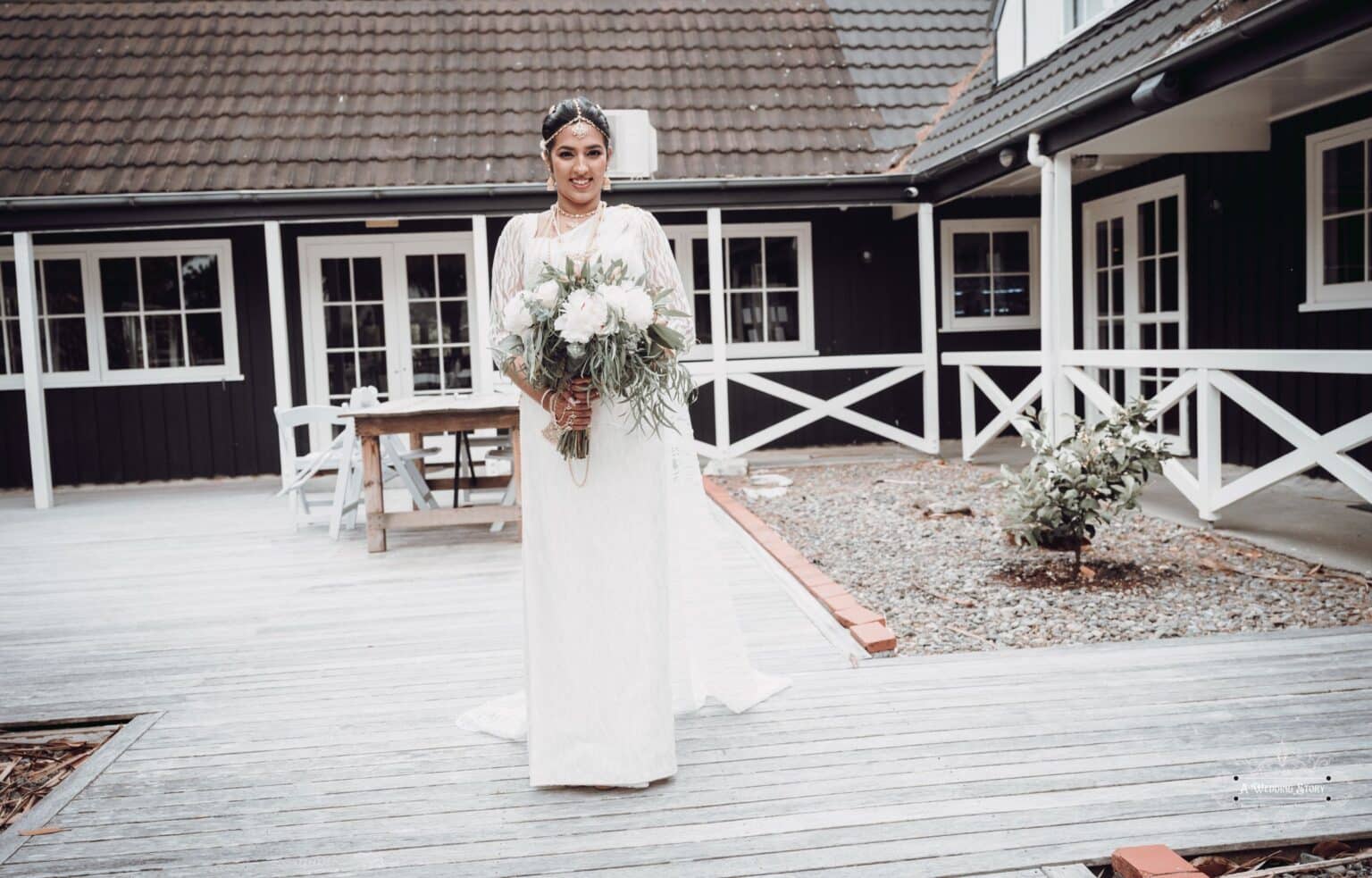 A stunning bride in a traditional Sri Lankan wedding saree holding a bouquet, standing on a rustic wooden deck at The Lodge, Wellington