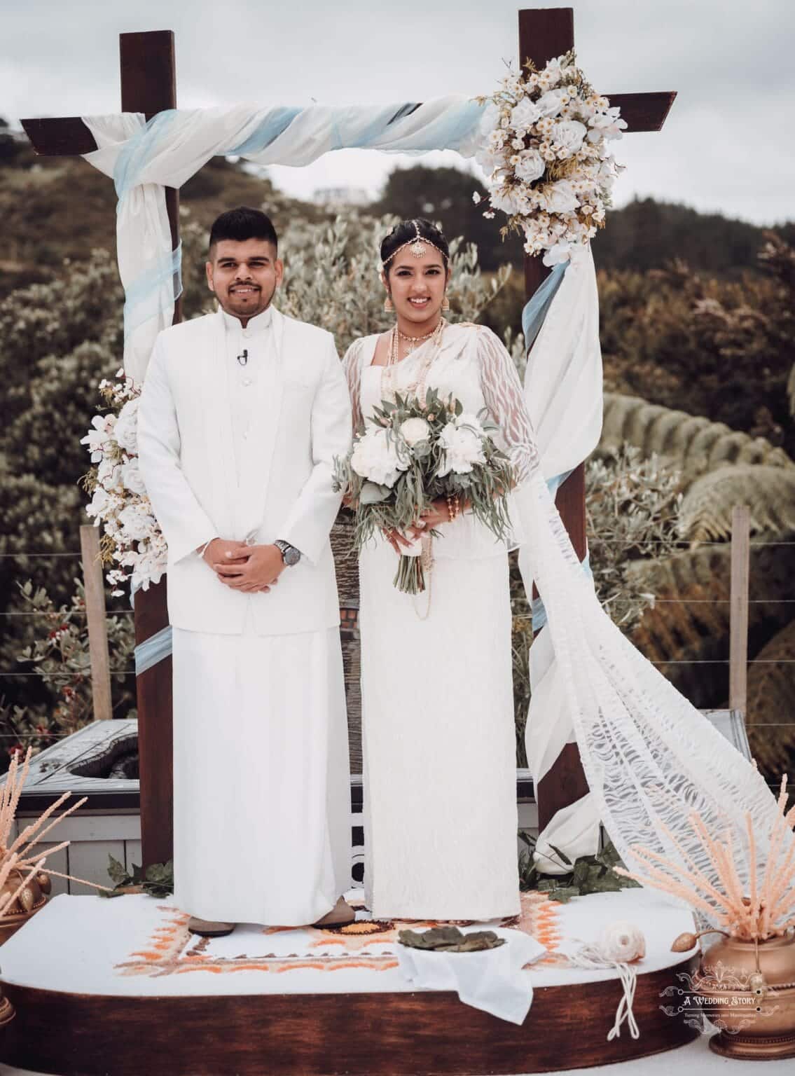 A newlywed couple stands joyfully at their wedding ceremony stage at The Lodge, Pauatahanui Inlet, surrounded by floral decorations and a scenic natural backdrop