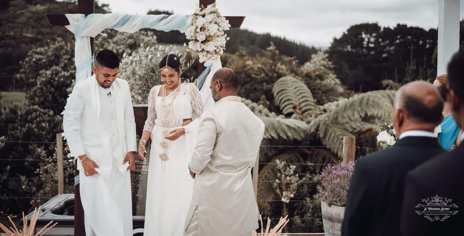 Bride and groom smiling during their wedding ceremony at The Lodge, Pauatahanui Inlet, with a floral-decorated cross in the background