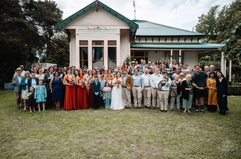 Bride and groom stand at the center of a large wedding group portrait in front of a charming countryside estate