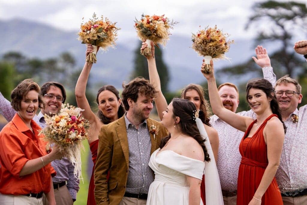 The bride and groom share a loving gaze as their wedding party raises bouquets in celebration with mountains in the background