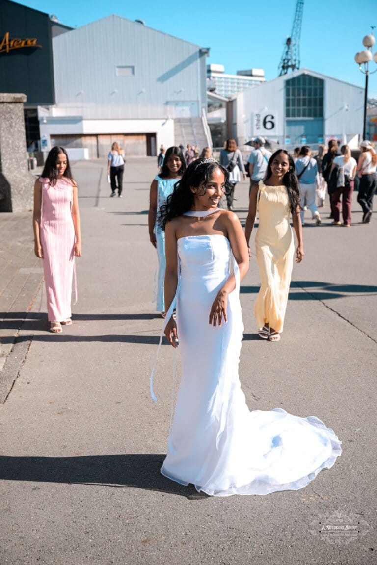 A bride in a white wedding gown walks with her bridesmaids in pastel dresses at the Wellington waterfront, smiling joyfully