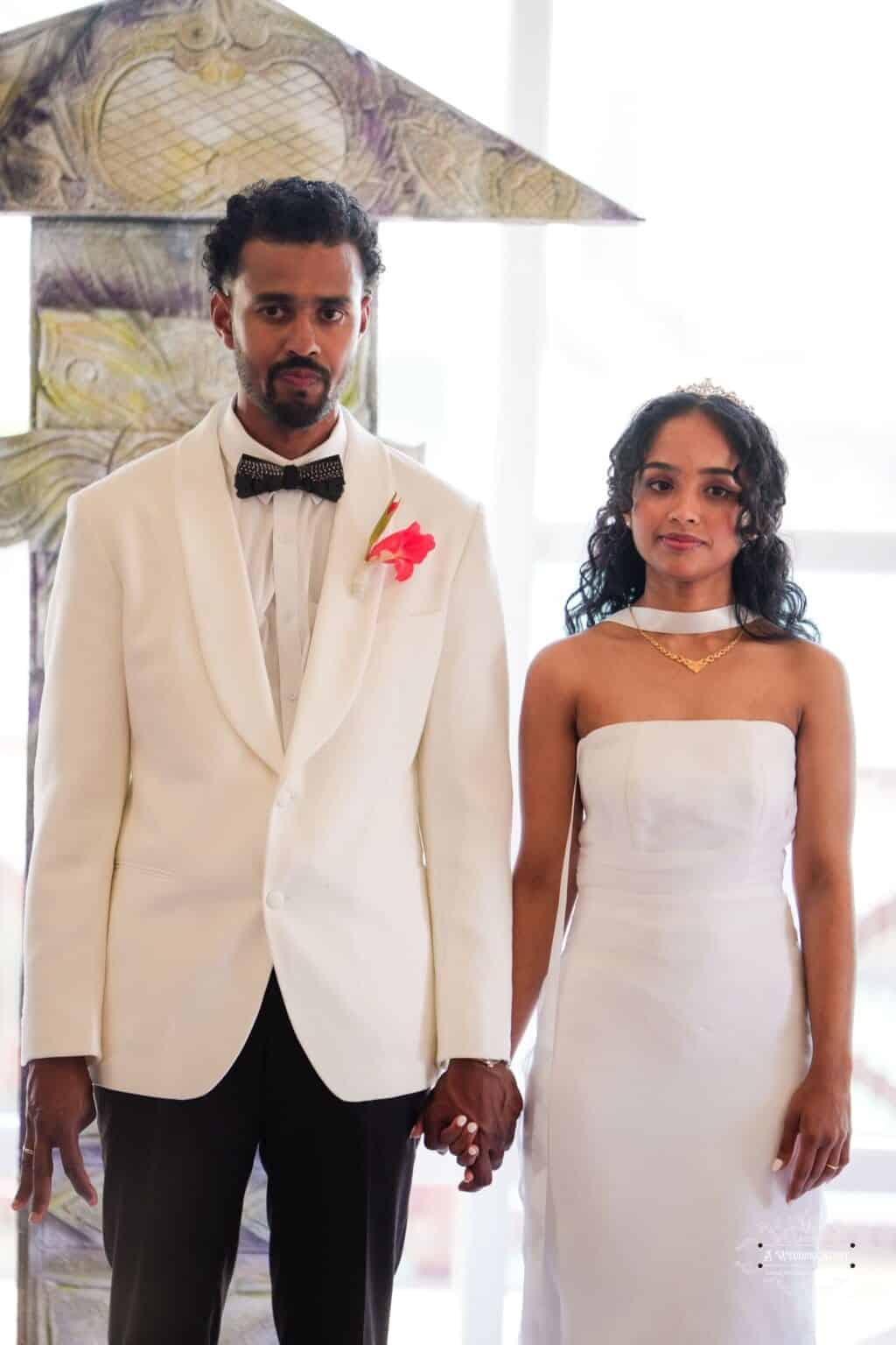 A newlywed couple holding hands during their wedding ceremony, dressed elegantly in white attire, with a traditional backdrop behind them