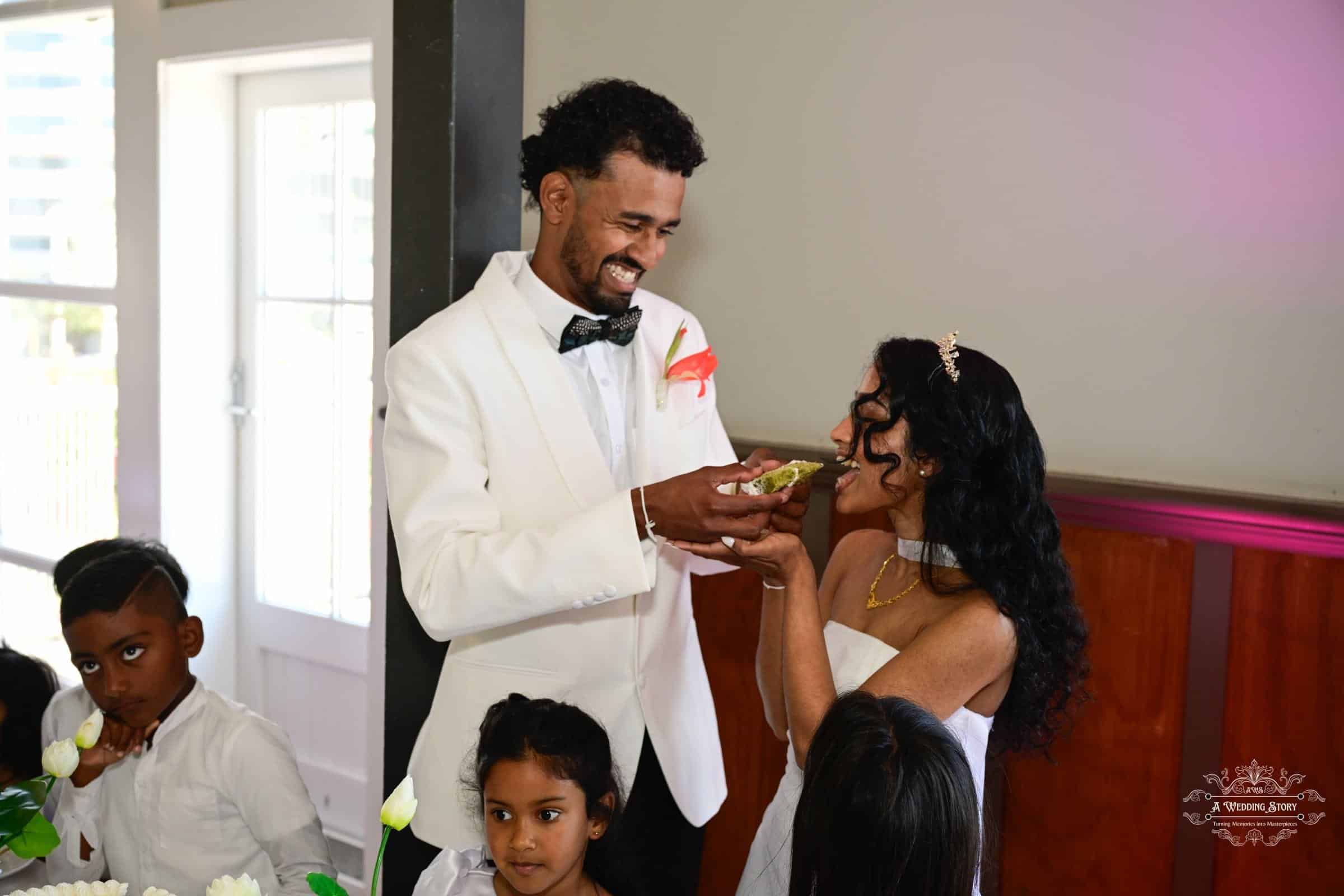 Groom playfully feeding the bride a traditional sweet during their wedding celebration, both smiling with joy