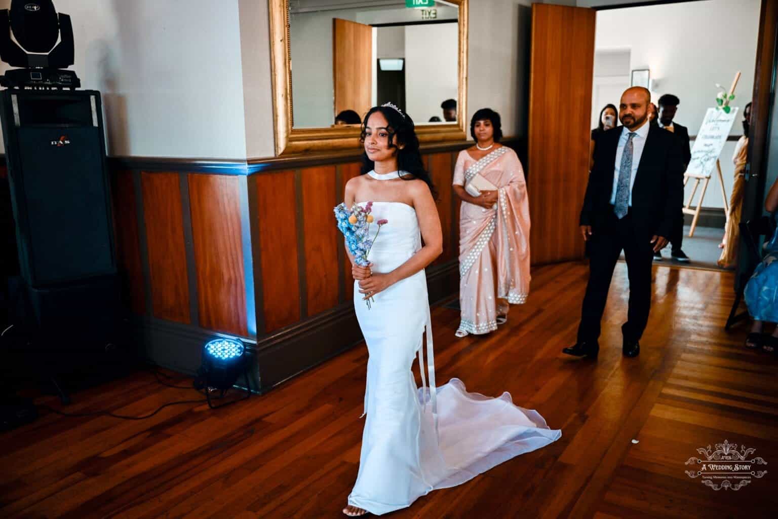 A bride in a stunning white gown walks down the aisle holding a bouquet, accompanied by family members in traditional attire at a wedding in Wellington, New Zealand