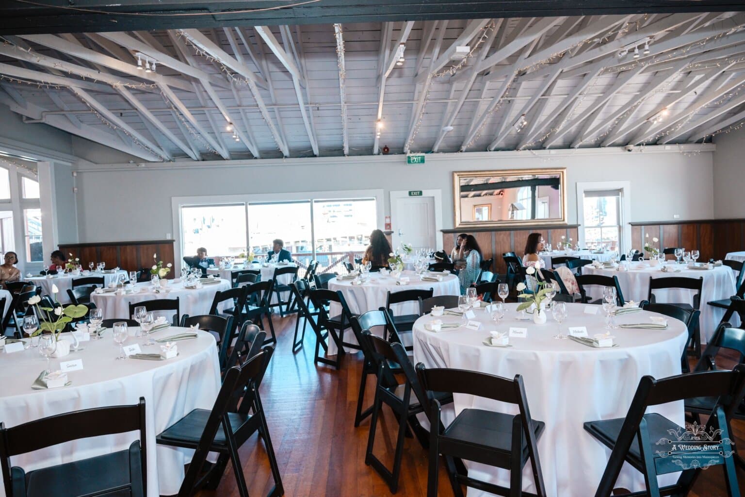 A beautifully decorated wedding reception venue with round tables, white tablecloths, floral centerpieces, and black chairs, set in a modern space with large windows