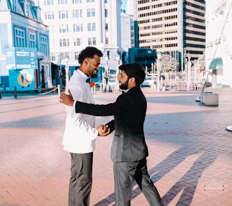 The groom in a white tuxedo and his best man in a black suit share a heartfelt moment in an urban plaza