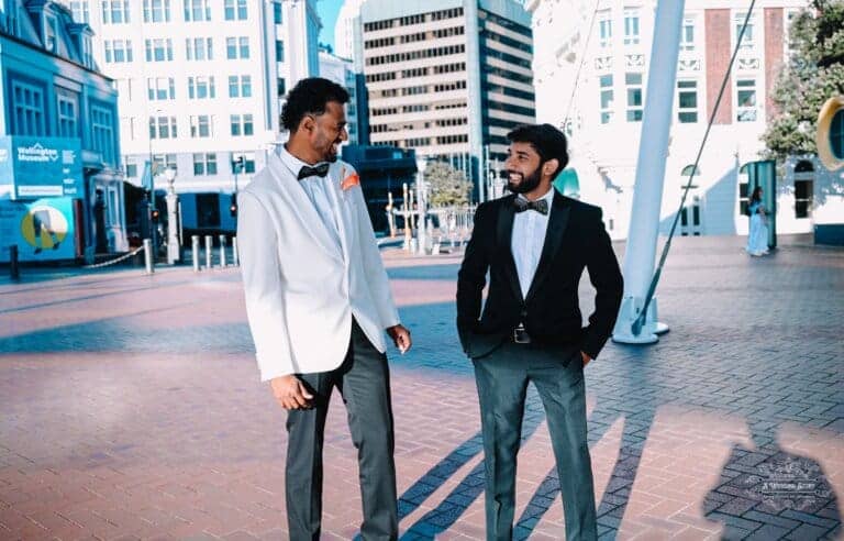 The groom in a white tuxedo and his best man in a black suit exchange smiles in a modern city square