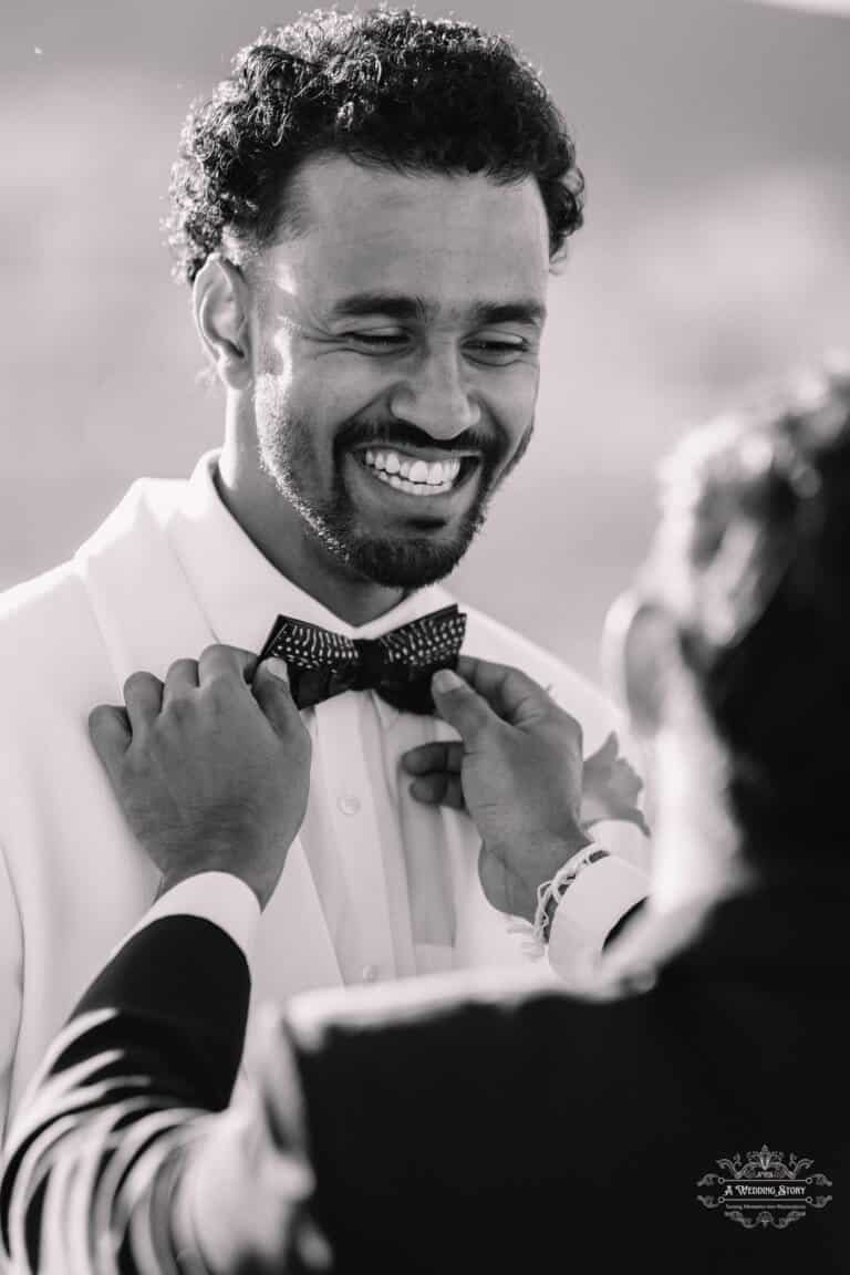 A groom smiling warmly as his best man adjusts his bow tie before the wedding ceremony