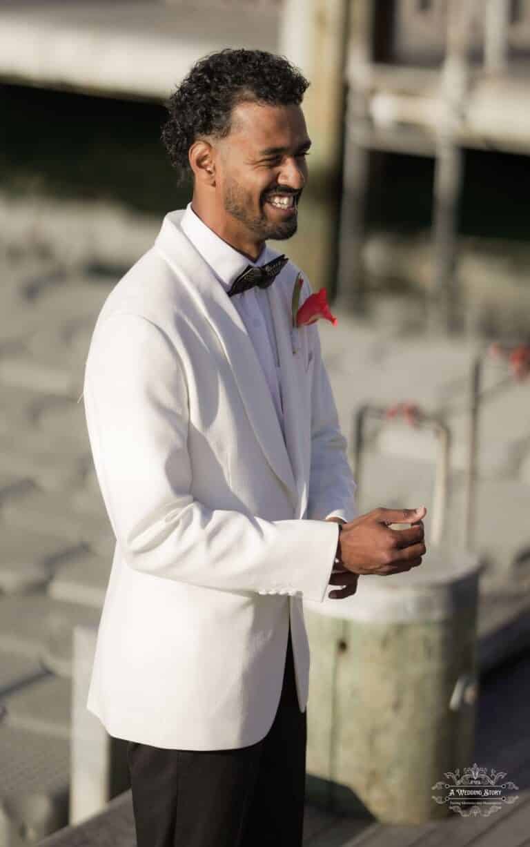 A groom in a white tuxedo smiles warmly while adjusting his cuff at the Wellington dock