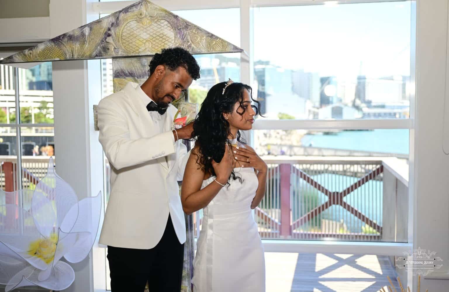 A groom lovingly places a necklace on his bride during their wedding ceremony, symbolizing love and commitment, with a scenic Wellington waterfront view in the background