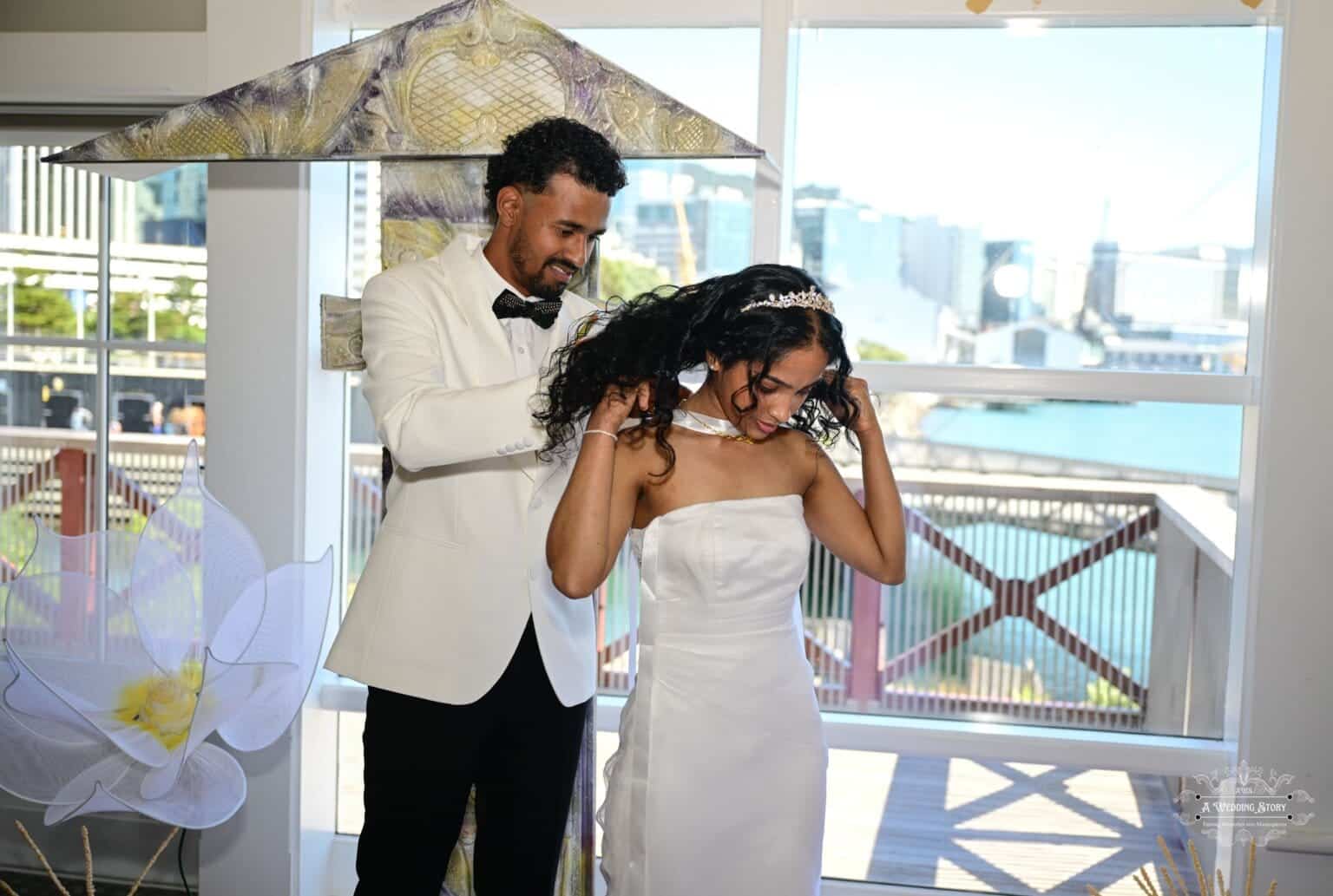 A groom gently places a necklace on his bride during their wedding ceremony, symbolizing love and commitment, with a picturesque Wellington waterfront in the background