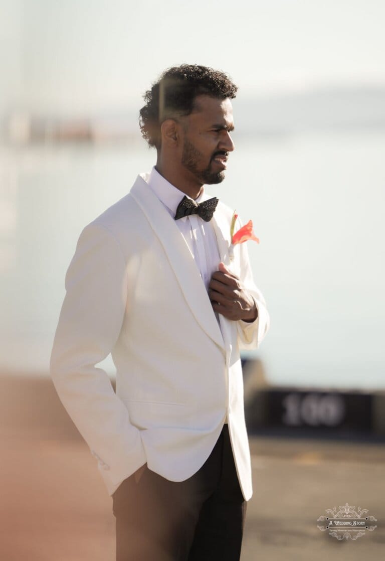 A groom in a white tuxedo with a bow tie and boutonnière stands by the waterfront, looking towards the horizon in soft golden light