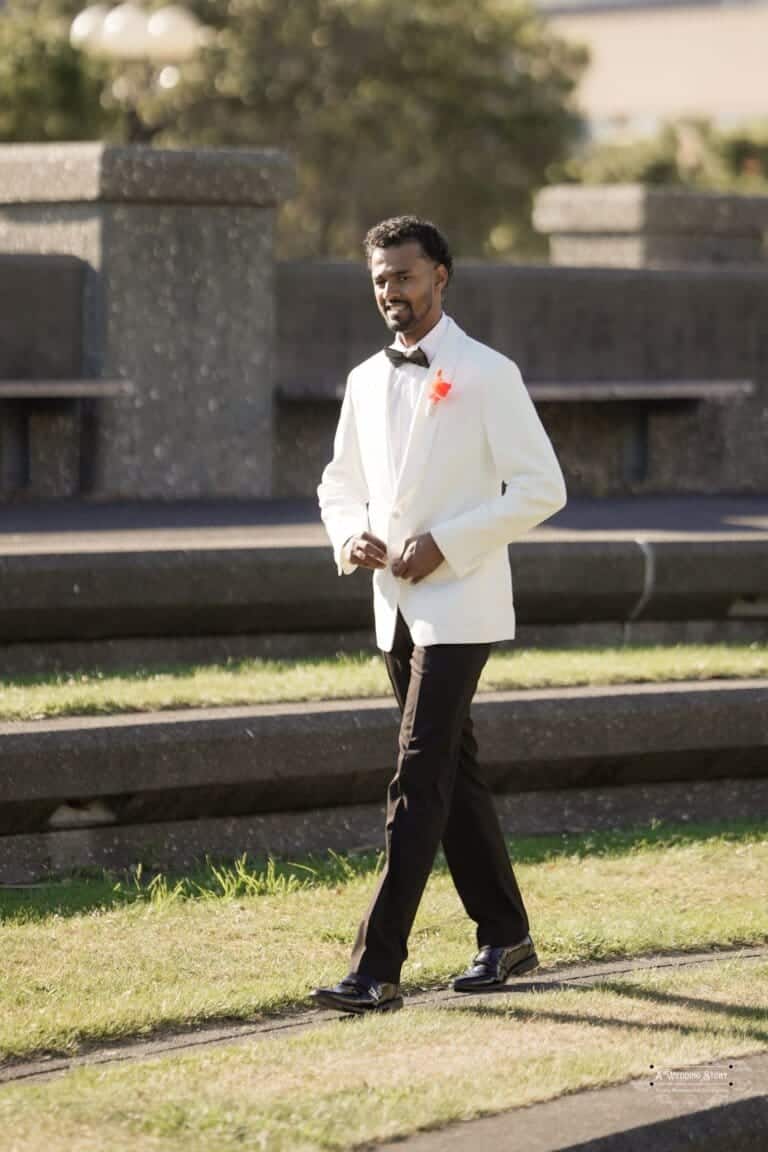 A groom in a white tuxedo and bow tie walks through a sunlit outdoor venue, radiating confidence and joy