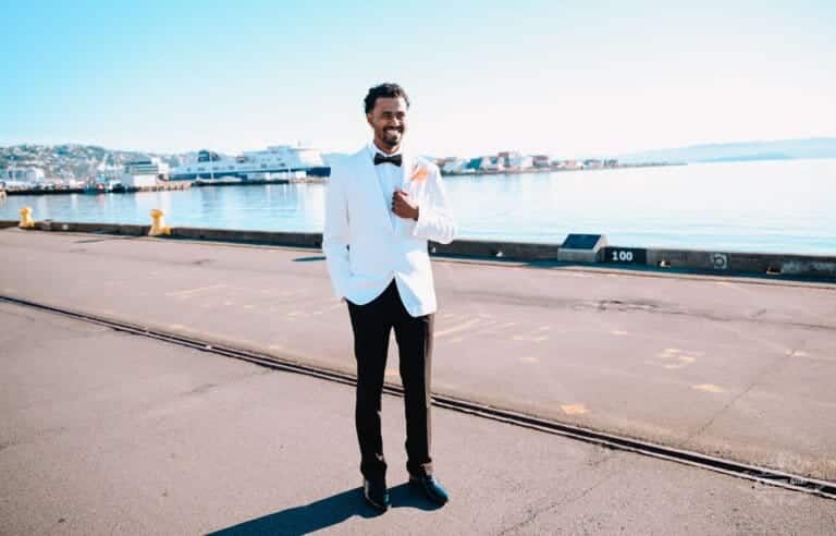 A groom in a white tuxedo stands confidently by the waterfront, smiling on his wedding day