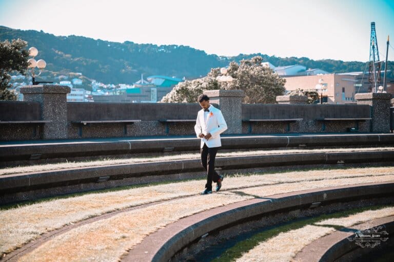 A groom in a white tuxedo walks through an outdoor amphitheater, adjusting his suit before the wedding