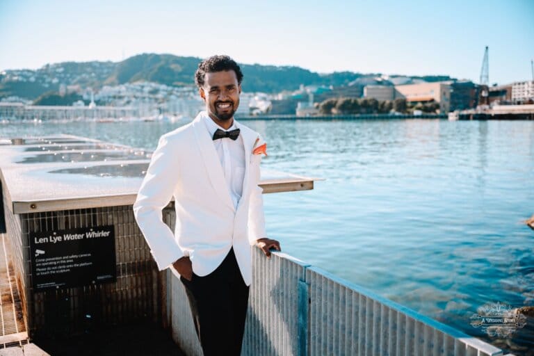 The groom in a white tuxedo smiles confidently while posing near the waterfront with Wellington cityscape in the background