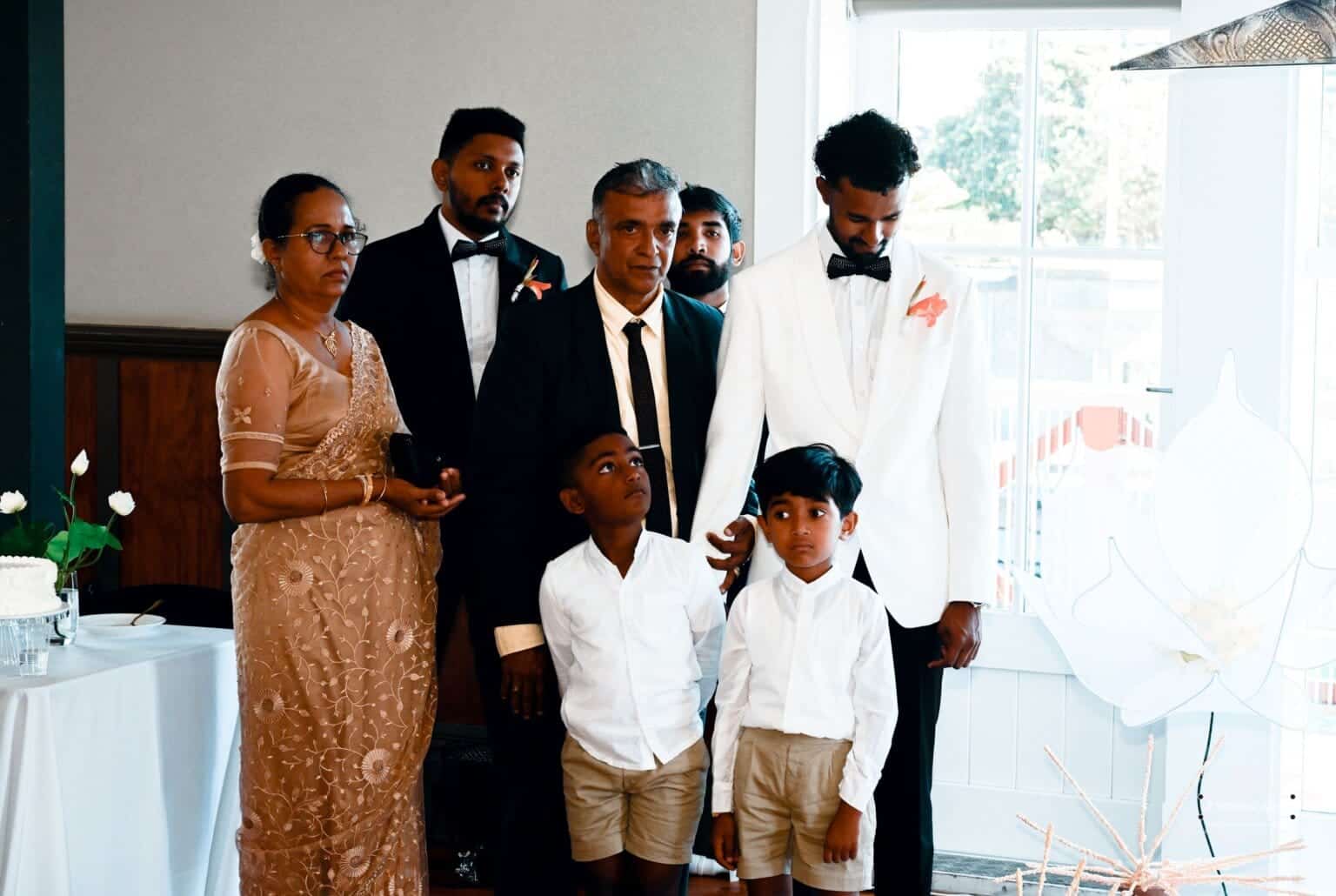 A groom in a white suit stands with his family, including two young boys and elegantly dressed relatives, during a heartfelt wedding moment in Wellington, New Zealand