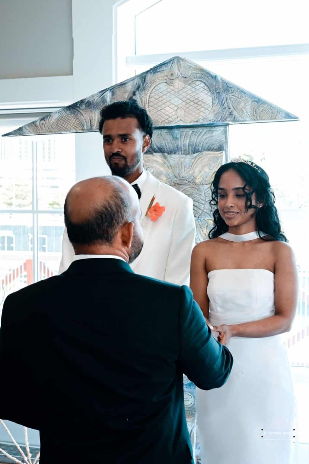 A newlywed couple stands together during their wedding ceremony, holding hands and receiving blessings from an elder in Wellington, New Zealand