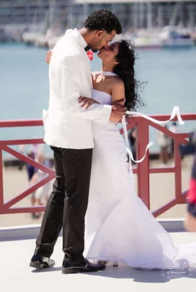 A newlywed couple shares a passionate kiss against the scenic waterfront, with the bride’s flowing ribbons adding a dreamy touch