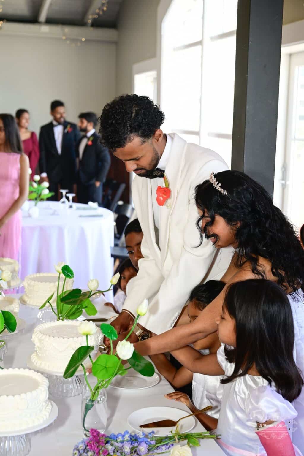 The bride and groom share a joyful moment cutting their wedding cake with children, surrounded by family and elegant decor at a Wellington wedding