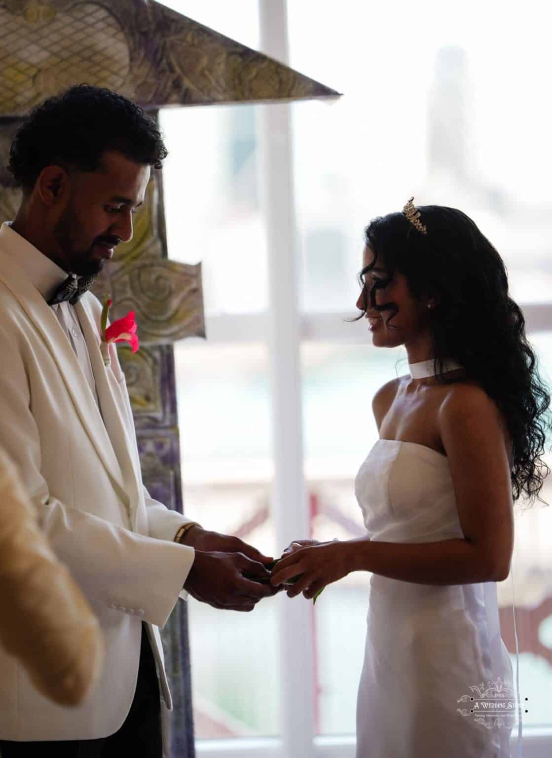 Bride and groom exchanging wedding rings in an intimate ceremony, with the groom in a white suit and the bride in a white gown with a tiara