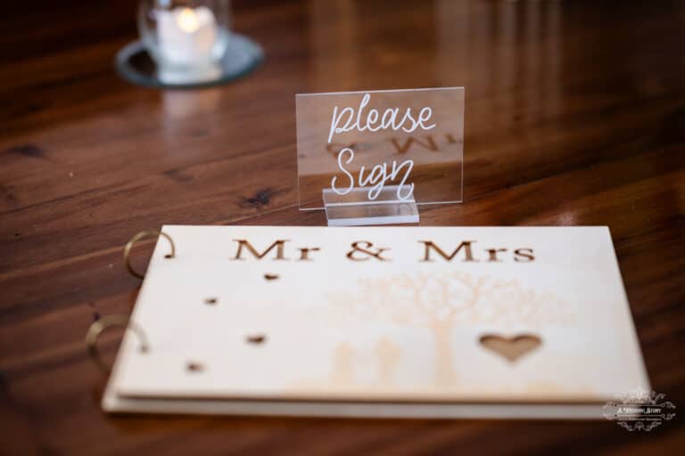 Rustic Mr & Mrs wedding guestbook with transparent “Please Sign” display on wooden table at The Lodge, Pāuatahanui
